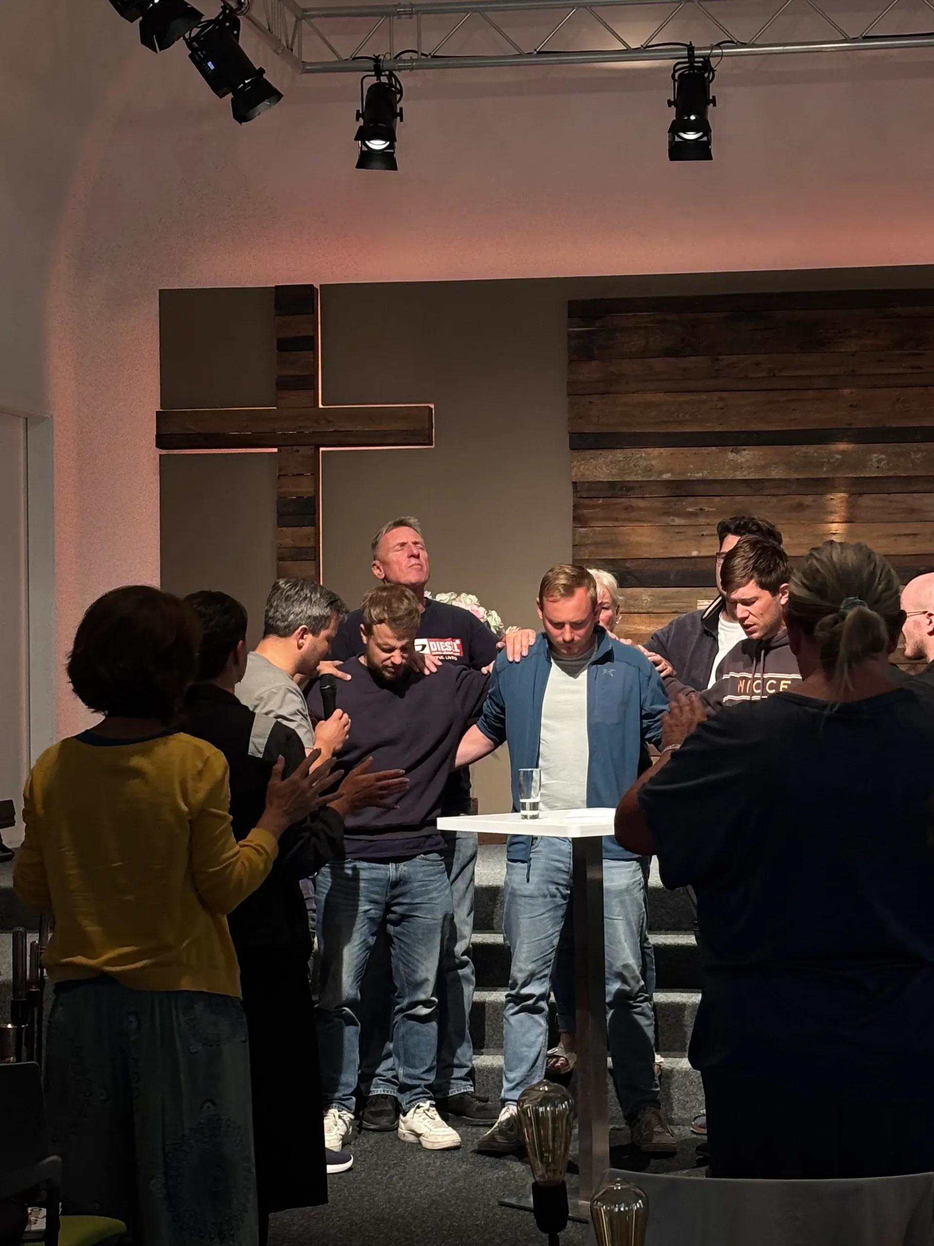 A group of people gathered around two men with heads bowed, placing hands on them in prayer in front of a wooden cross mounted on the wall.