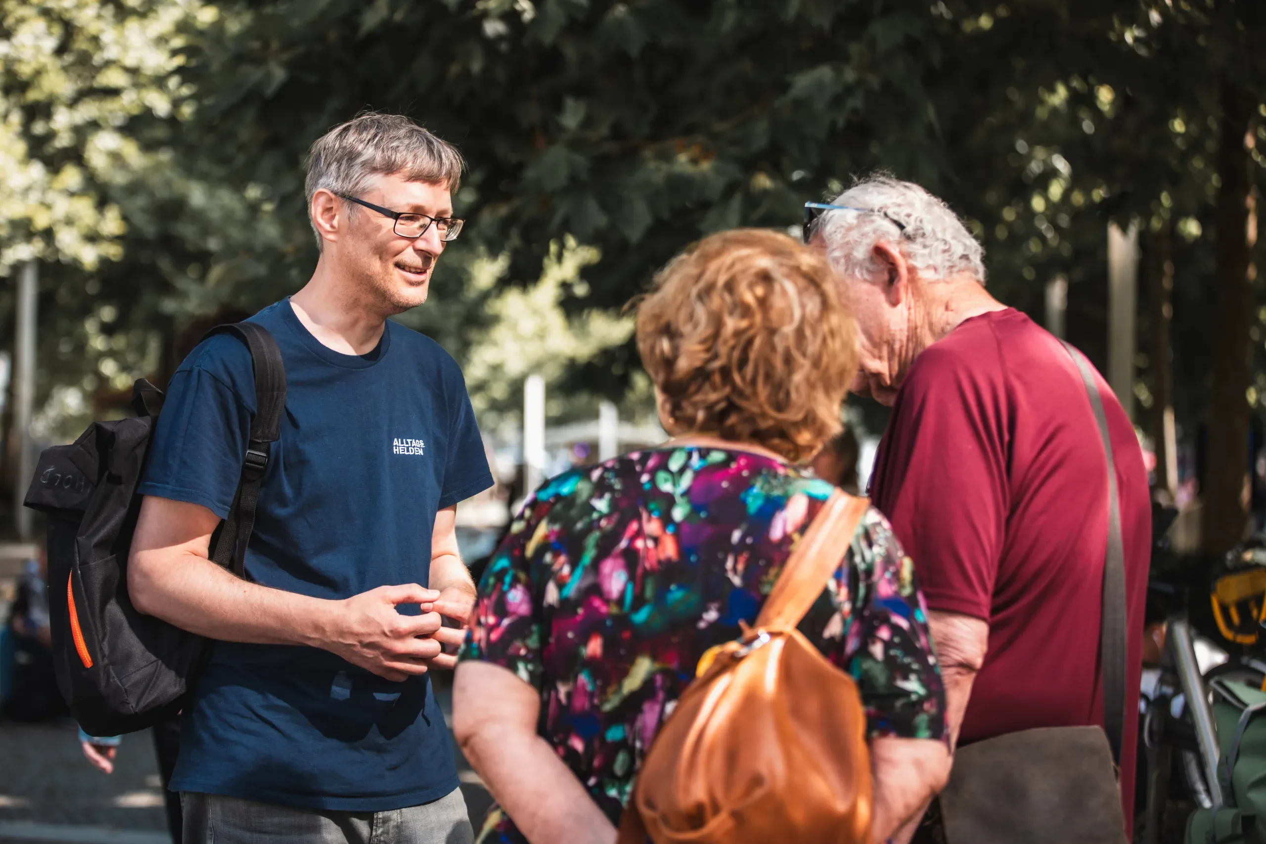 A man in a navy "Alltags Helden" T-shirt and glasses talks with an older couple outdoors among trees.
