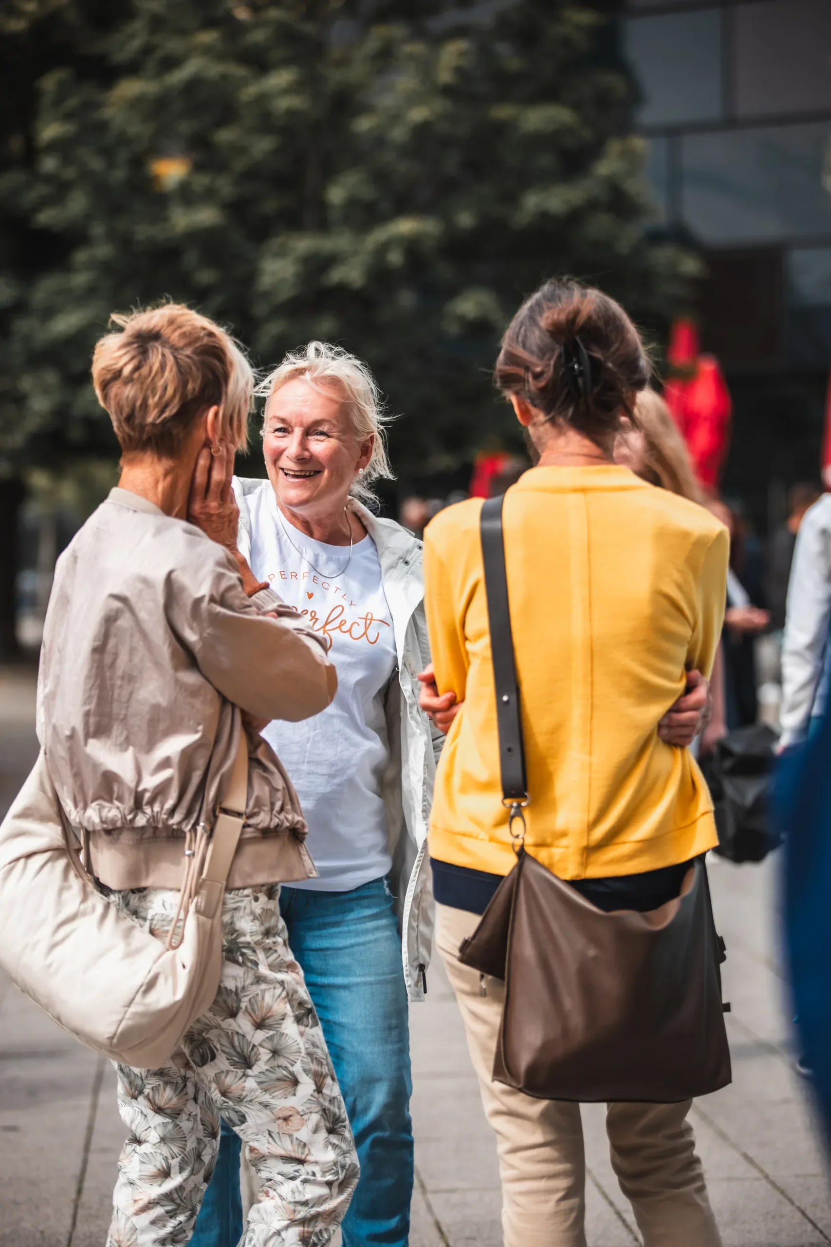 A smiling woman in a "Perfectly Perfect" t-shirt talks with two other women on a tree-lined sidewalk.