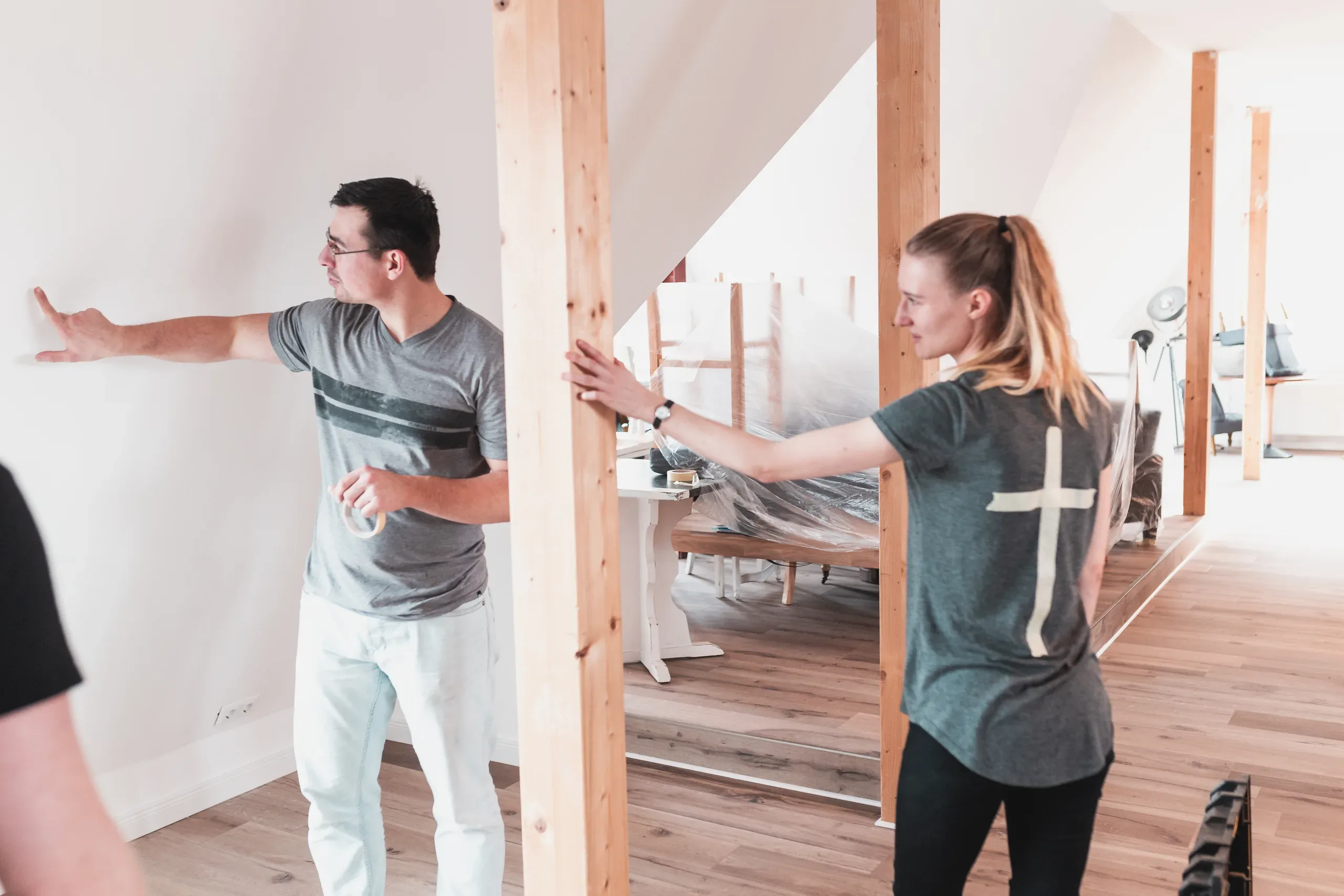 A man holding a roll of masking tape and a woman with a cross on her shirt stand on either side of a wooden beam in a room with furniture covered in plastic sheeting.