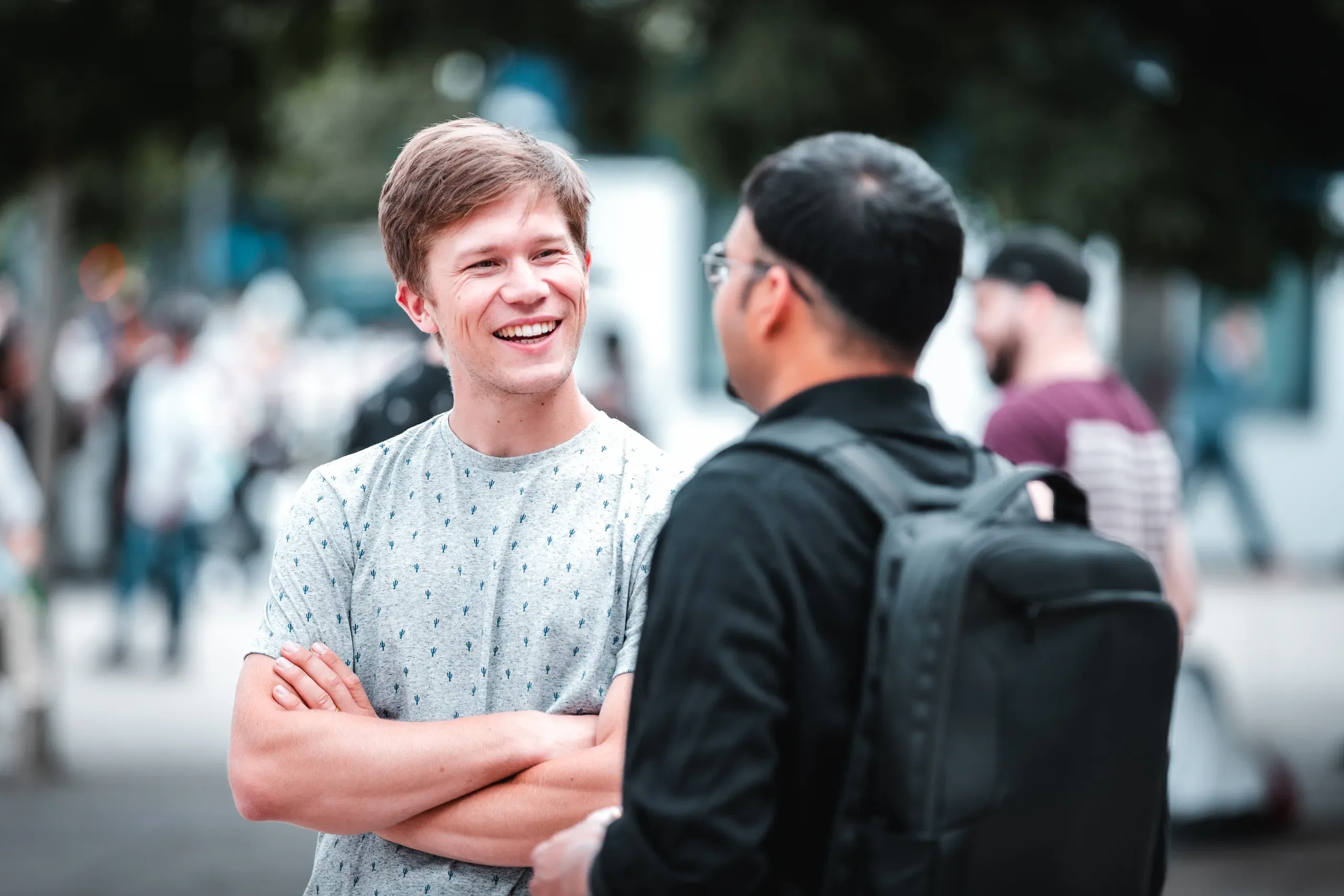 A smiling man with crossed arms in a cactus-print t-shirt talks with another man carrying a black backpack on a crowded street.