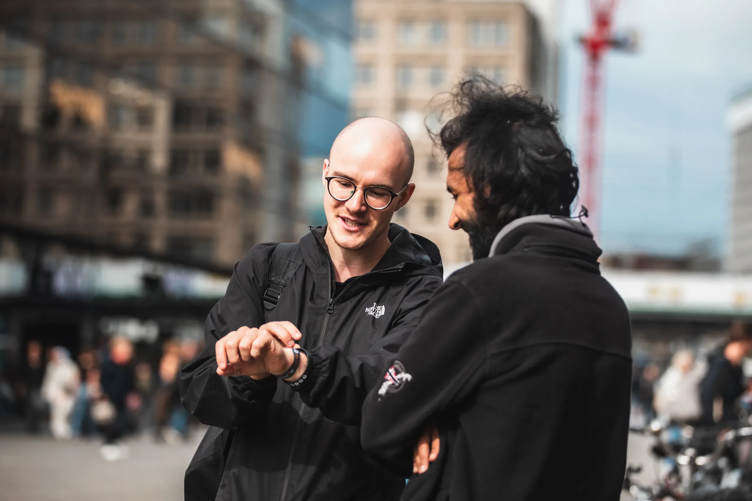 Two men in black jackets look at a wristwatch on a busy city street.